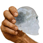 Handgearbeiteter Kristall-Totenkopf aus echtem Bergkristall – gehalten in kräftiger, leicht verschmutzter Männerhand auf weissem Hintergrund. Glasklarer Kristallschädel, Symbol für Stärke, Energie, Klarheit und nordische Handwerkskunst.