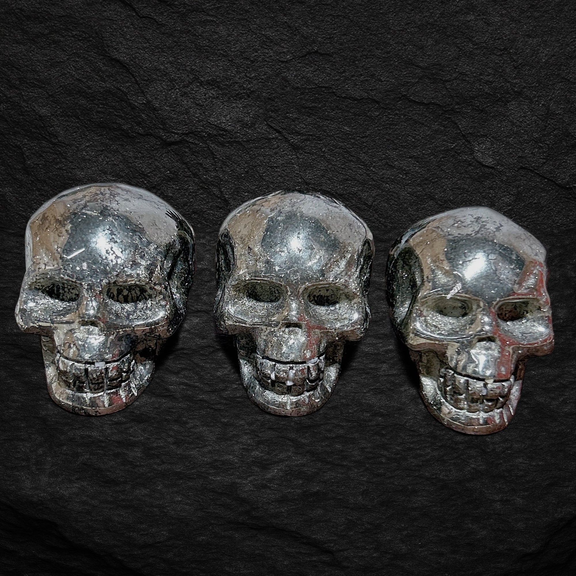Drei echte Pyrit-Totenköpfe auf dunklem Steinuntergrund – glänzende Wikinger-Schädel aus massivem Pyrit, Symbol für Stärke, Mut, Schutz und Handwerkskunst.