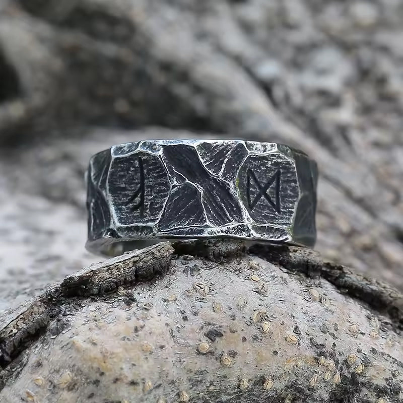 Runen Ring aus Edelstahl mit mehreren eingravierten Runen – liegend auf Felsen im Wikinger-Stil – strukturierte, dunkle Oberfläche mit antiker Optik – symbolträchtiger Ring für Stärke und Schutz, hochwertiger Wikinger Runenschmuck von NORDSKULLS.