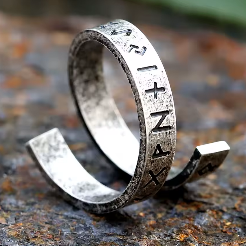 Spiralförmiger Wikinger-Ring aus antikem Silbermetall mit tief eingravierten Runen. Die schwarzen Runensymbole verlaufen gleichmässig entlang der leicht geöffneten Ringschiene. Der Ring liegt auf einer rauen, rostfarbenen Steinoberfläche – im rustikalen, nordischen Stil fotografiert.