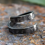 Dunkler Wikinger-Ring in Spiralform mit eingravierten Runen, gefertigt aus gealtertem Metall. Die feinen Runensymbole verlaufen über beide Ringhälften, die sich spiralförmig umeinander winden. Der Ring liegt auf einer rauen, rostfarbenen Oberfläche – detailreich, rustikal und im authentischen Wikinger-Stil fotografiert.