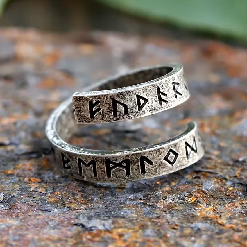 Antiker Wikinger-Ring aus silberfarbenem Metall mit spiralförmigem Design und tief eingravierten Runen. Die schwarzen Runensymbole verlaufen über zwei verwitterte Ringbänder, die sich kunstvoll umeinander winden. Der Ring liegt auf einem rostfarbenen, steinartigen Untergrund – rustikal, naturverbunden und im authentischen Wikinger-Stil.