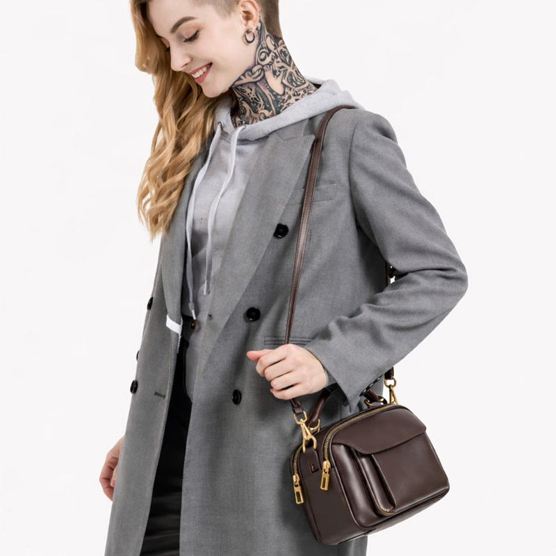 Damen Echtleder Handtasche in der Farbe Coffee als Schultertasche getragen, hochwertiges echtes Leder mit goldenen Metallbeschlägen, modernes Damen-Model im grauen Mantel vor weissem Hintergrund.