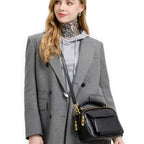 Damen Echtleder Handtasche in Schwarz als Schultertasche getragen, hochwertiges echtes Leder, goldene Metallbeschläge, modernes Damen-Model mit grauem Mantel vor weissem Hintergrund.